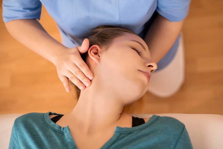 Person gently stretching a sore shoulder in a bright home beside a chair and heating pad